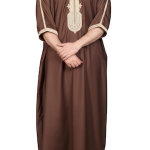 May include: A brown, long-sleeved garment with beige embroidery around the neckline and sleeves. The traditional attire features a loose fit and is suitable for various occasions. The garment is worn by a person.