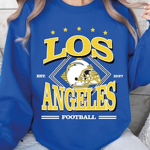 Puede incluir: Sudadera azul real con el texto "LOS ANGELES FOOTBALL" en amarillo y blanco, y un gráfico de casco de fútbol. La sudadera tiene cuello redondo y mangas largas. También se incluye el texto "EST. 1937".