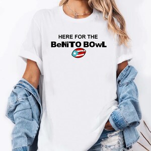 May include: White t-shirt with the text "HERE FOR THE BeNiTO BOWL" in black, with a football graphic featuring the Puerto Rican flag. The shirt is worn with a denim jacket.