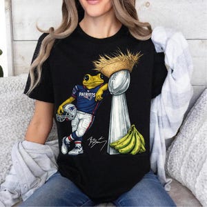 Puede incluir: Camiseta negra con una rana de dibujos animados con un uniforme de fútbol de los Patriots y un sombrero de paja, apoyada en un trofeo plateado. Un racimo de plátanos está al lado del trofeo. La rana sostiene un casco de fútbol.