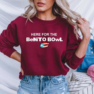 May include: A maroon sweatshirt with the text "HERE FOR THE BeNITO BOWL" in white. The word "BOWL" features a football graphic with the Puerto Rican flag. The sweatshirt is a casual garment.