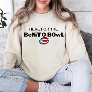 May include: Cream-colored sweatshirt with the text "HERE FOR THE BeNiTO BOWL" in black. A football graphic with the Puerto Rican flag is below the text. The sweatshirt is worn by a person.