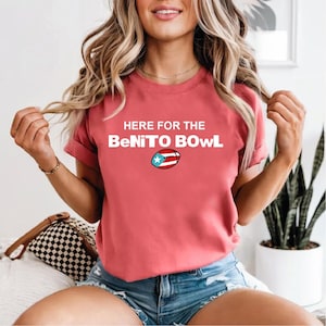 Puede incluir: Camiseta de color coral con el texto blanco "HERE FOR THE BeNITO BOWL". Un gráfico de fútbol con la bandera de Puerto Rico está debajo del texto. La camiseta es de manga corta.