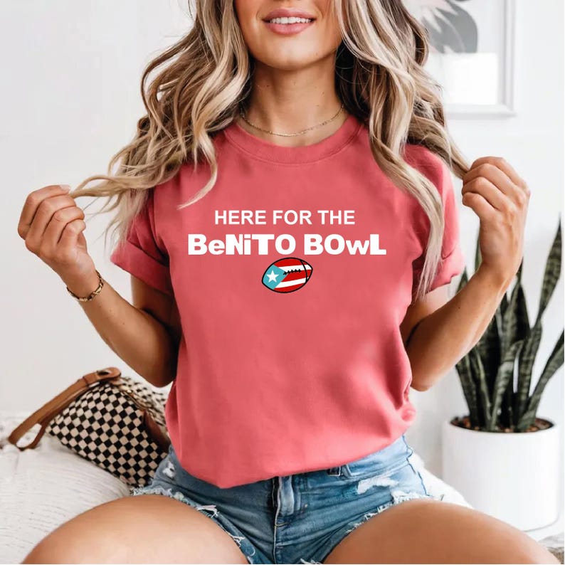 May include: A coral-colored t-shirt with the text "HERE FOR THE BeNITO BOWL" in white. A football graphic with the Puerto Rican flag is below the text. The shirt is short-sleeved.