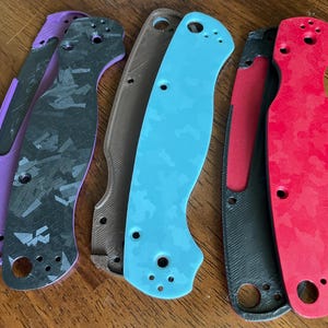 May include: A collection of knife handle scales in various colors, including purple, black, brown, teal, and red. The scales have a textured pattern and several holes for attachment. The scales are designed for a specific knife model.