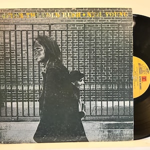 May include: Album cover for Neil Young's "After the Gold Rush" vinyl record. The cover features a sepia-toned image of a man and woman walking in front of a brick wall and a metal fence. The album title and artist's name are at the top.