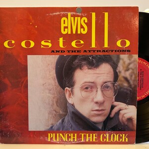May include: A red and yellow vinyl record album cover featuring Elvis Costello and The Attractions. The album title is "Punch The Clock".
