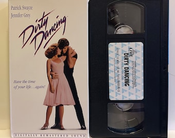 May include: A VHS tape of the movie Dirty Dancing, featuring Patrick Swayze and Jennifer Grey on the cover. The cover is white with purple text and a photo of the actors dancing. The text on the tape reads "Live Dirty Dancing (Original Version)" and "Approx. 105 Mins./Cat. No. 16013/Rated PG-13".