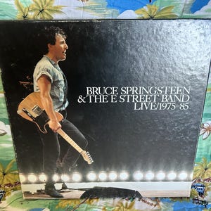 May include: A vintage album cover featuring Bruce Springsteen & The E Street Band. The image shows Springsteen holding a guitar on stage, with the album title "LIVE/1975-85" in white text. The background is black with stage lights.