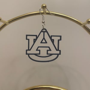 May include: A pair of black and white earrings featuring the letter 'A' with a circle around it. The earrings are hanging from a gold stand.