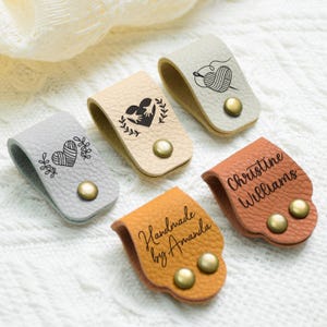 May include: A collection of leather yarn labels in various colors, including gray, tan, and brown. Each label features a unique design or text, such as a heart, yarn and needle, or the name "Christine Williams". They are secured with brass-colored snaps.