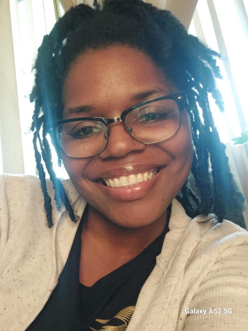 K&ouml;nnte beinhalten: Ein Nahaufnahmeportr&auml;t einer Person mit dunkelbrauner Haut, die eine schwarze Brille und einen hellbeigen Pullover &uuml;ber einem schwarzen T-Shirt tr&auml;gt. Die Person hat lange, dunkelbraune Dreadlocks und l&auml;chelt. Das Bild ist aus der Selfie-Perspektive aufgenommen und der Text "Galaxy A52 5G" ist in der rechten unteren Ecke sichtbar.