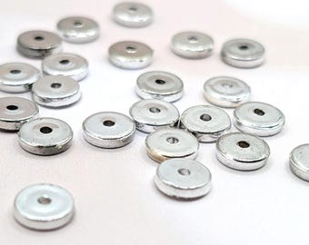 Könnte beinhalten: Eine Ansammlung kleiner, silberfarbener, flacher, runder Metallperlen mit einem Loch in der Mitte. Die Perlen sind auf einer weißen Oberfläche verstreut. Geeignet für die Schmuckherstellung und andere Bastelprojekte.
