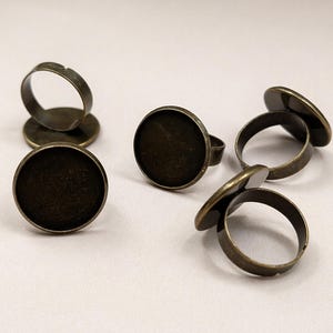 Può includere: Diversi supporti per anelli regolabili color bronzo antico con castone rotondo. Gli anelli sono sparsi su una superficie bianca. I castoni sono progettati per contenere un inserto rotondo, perfetti per creare gioielli personalizzati.
