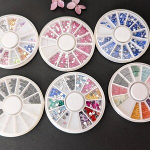 Peut inclure: Six organisateurs circulaires blancs pour nail art remplis de divers strass et embellissements colorés. Les strass sont dans les tons de rose, bleu, vert et or. Les organisateurs sont disposés sur une surface sombre.