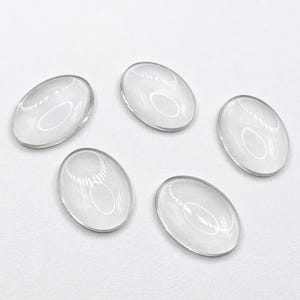 Op de afbeelding: Vijf heldere, ovale glazen cabochons. Elke cabochon is transparant met een licht afgeronde bovenkant en een platte bodem. De cabochons zijn gerangschikt op een wit oppervlak en reflecteren het licht.