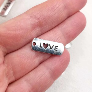 Peut inclure: Un petit charme rectangulaire argenté avec le mot "LOVE" découpé et une découpe en forme de cœur au centre. Le charme a un petit trou à une extrémité, probablement pour l'enfilage. Le charme est tenu dans une main.