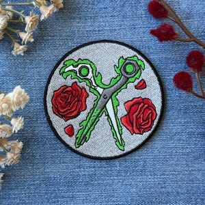 May include: Embroidered patch featuring crossed scissors with green handles and a gray blade, surrounded by two red roses and petals on a gray background. The patch has a black border and is set against a blue denim backdrop.