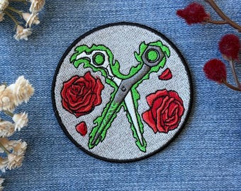 May include: Embroidered patch featuring crossed scissors with green handles and a gray blade, surrounded by two red roses and petals on a gray background. The patch has a black border and is set against a blue denim backdrop.