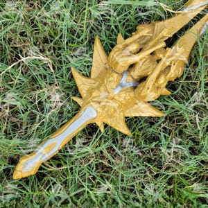 May include: A gold-colored sword with a silver blade and intricate details. The sword has a decorative handle with a lion's head and a crown. The sword is lying on a bed of green grass.