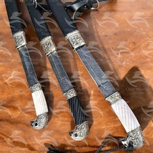 May include: Three ornate swords with black scabbards and silver accents. The swords have different coloured handles: white, black, and a combination of black and white. The swords are displayed on a brown wooden surface.