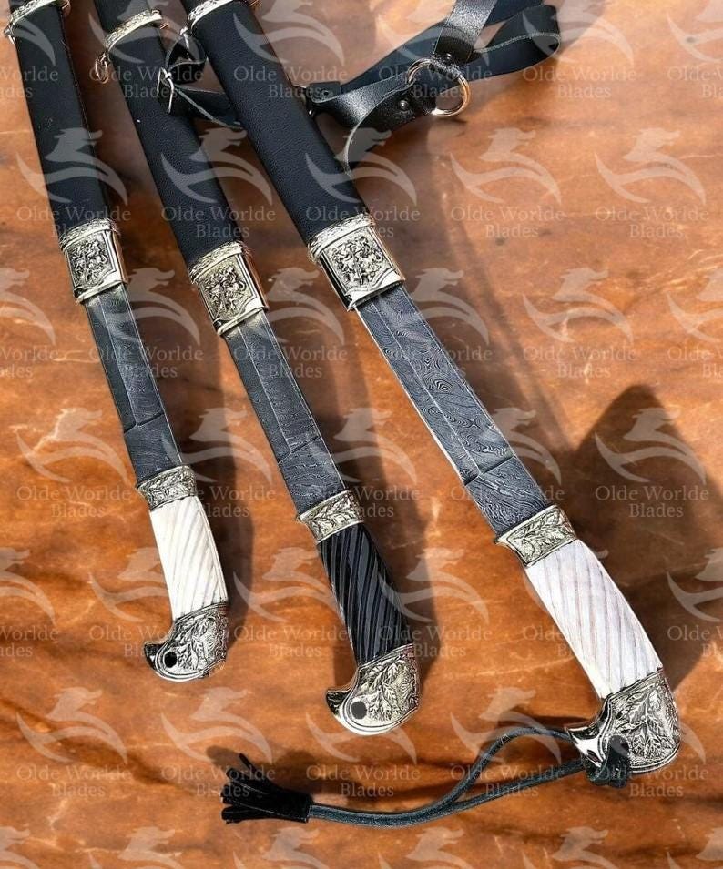 May include: Three ornate swords with black scabbards and silver accents. The swords have different coloured handles: white, black, and a combination of black and white. The swords are displayed on a brown wooden surface.