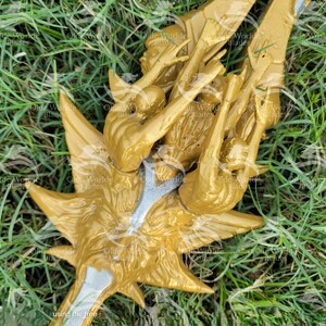 May include: A gold-colored metal sculpture of a mythical creature with a long, pointed tail and wings. The sculpture is detailed with intricate carvings and is set against a backdrop of green grass.
