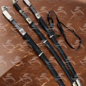 May include: Three black leather-wrapped swords with silver accents and ornate handles. The swords are displayed on a brown surface with a black leather strap.