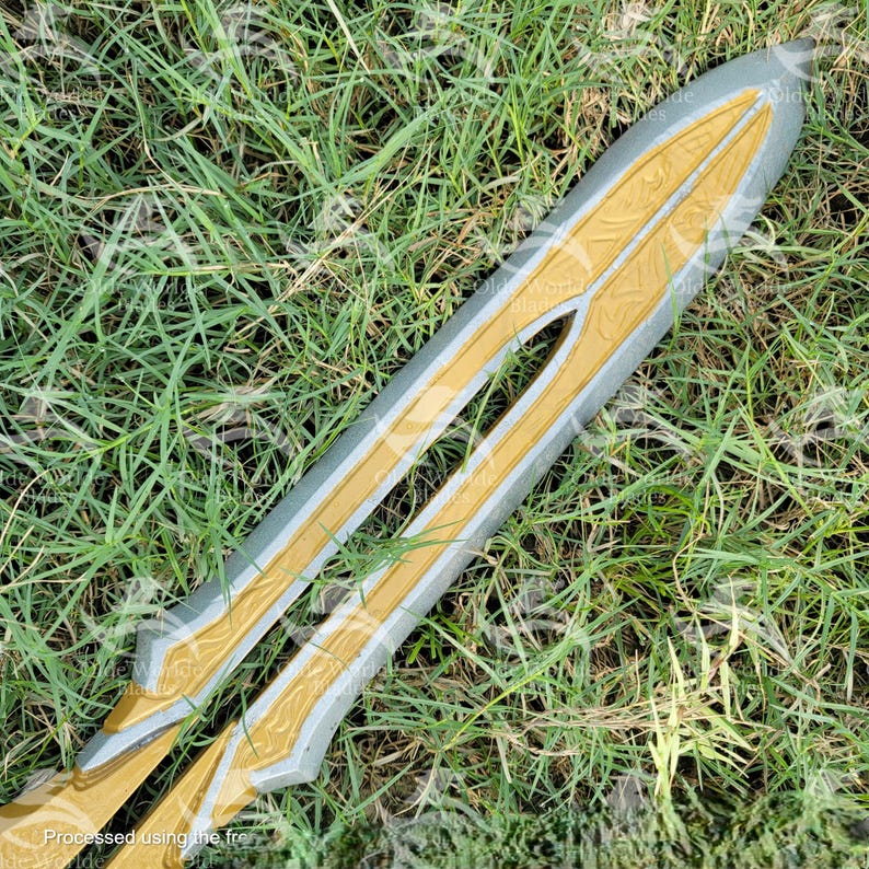 May include: A silver and gold sword with intricate details on the blade. The sword is lying on a bed of green grass.