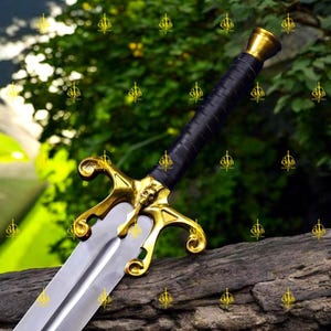 Può includere: Una spada decorativa con un'elsa dorata con un design a teschio e un'impugnatura avvolta in pelle nera. La lama argentata poggia su una superficie di legno, con uno sfondo verde sfocato.
