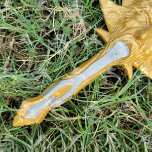 May include: A gold and silver decorative sword with a detailed handle. The sword is lying on a bed of green grass.