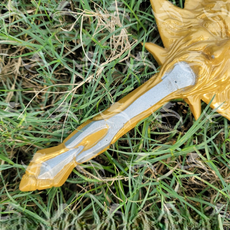 May include: A gold and silver decorative sword with a detailed handle. The sword is lying on a bed of green grass.