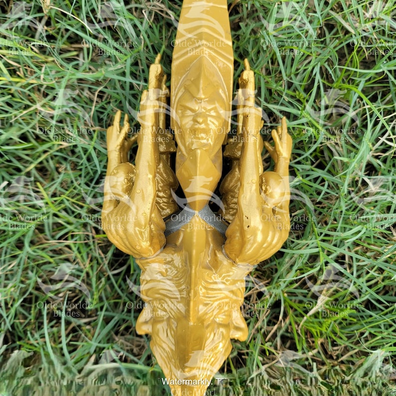 May include: A gold-colored, three-dimensional sculpture of a creature with multiple arms and a human-like face. The sculpture is set against a background of green grass.