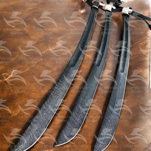 May include: Three black swords with intricate patterns on the blades. The swords are arranged in a fan shape on a brown wooden surface.