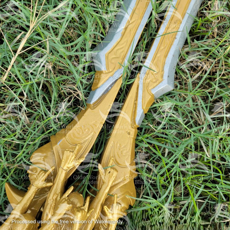 May include: A gold and silver sword with intricate details, lying on a bed of green grass. The sword has a sharp point and a double-edged blade.