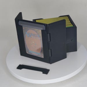以下が含まれることがあります： 黒いプラスチック製のカード収納ボックス。ヒンジ付きの蓋があり、緑色のカードの束が入っています。ボックスには、Magic: The Gatheringカードを表示する透明なプラスチック製の窓があります。別の黒いプラスチック片が前にあります。