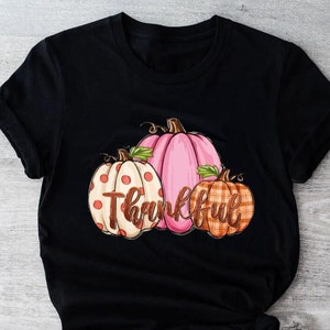 May include: Black t-shirt with a graphic of three pumpkins. The pumpkins are pink, white with red polka dots, and orange with a plaid pattern. The text "Thankful" is written in brown cursive letters.