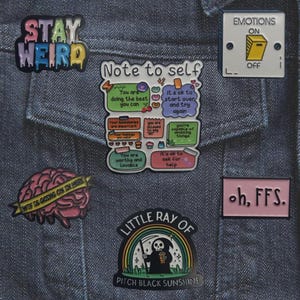 May include: A collection of enamel pins on a denim jacket. Pins include phrases like "Stay Weird," "Note to Self," and "Emotions On." Other pins feature a brain, a grim reaper under a rainbow, and the phrase "oh, FFS."