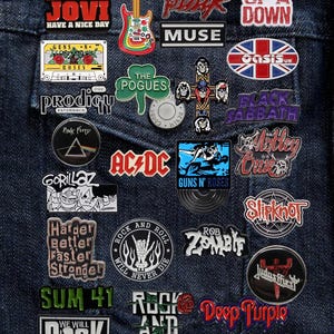 Puede incluir: Un conjunto de pines esmaltados coloridos con logotipos de bandas de rock como AC/DC, Guns N' Roses y Black Sabbath, fijados a una chaqueta vaquera. Los pines presentan colores vibrantes y diseños diversos, reflejando las identidades de las bandas.
