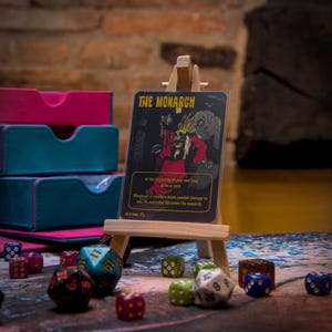 Könnte beinhalten: Eine schwarze und gelbe Karte mit dem Text "THE MONARCH" auf einem Holzstaffelei. Die Karte zeigt eine schwarze und rote Illustration einer skelettartigen Figur, die eine Krone trägt und einen Stab hält. Der Text auf der Karte lautet: "Zu Beginn deines Endschritts, ziehe eine Karte. Wann immer eine Kreatur Kampfschaden an dich verursacht, wird ihr Beherrscher zum Monarchen."