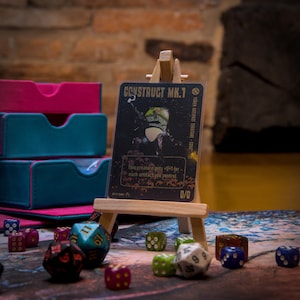 Può includere: Una carta da gioco con sfondo nero e testo dorato. La carta è intitolata "Construct MK.1" e presenta un robot con un casco verde. La carta è su un piccolo cavalletto di legno.