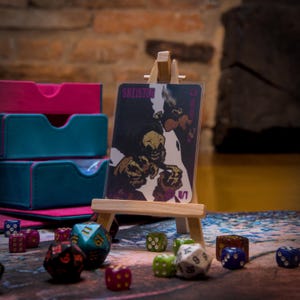 May include: A card game card with a purple and white illustration of a skeleton with the text "SKELETON" on the card. The card is on a small wooden easel and surrounded by colorful dice.