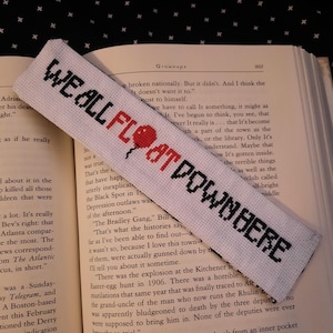 May include: A white cross-stitch bookmark with the phrase "WE ALL FLOAT DOWN HERE" in black and red lettering and a red balloon graphic. The bookmark is on the pages of an open book.