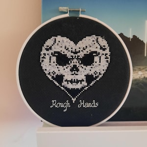 Könnte beinhalten: Schwarzer Stickrahmen mit einem weißen Totenkopf- und Knochen-Design in Form eines Herzens. Der Text "Rough Hands" ist unter dem Schädel gestickt.