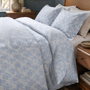 May include: A bed with a blue and white floral patterned duvet cover and matching pillowcases. The bedroom has a wooden nightstand, a woven headboard, and a patterned rug. A vase of flowers sits on the nightstand.