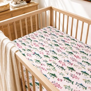 May include: A baby crib with a fitted sheet featuring a repeating pattern of colorful fish in shades of green, pink, and blue on a light pink background. The crib is made of light-colored wood.
