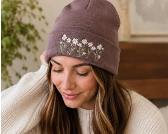 Puede incluir: Un gorro de punto color malva con un diseño bordado de flores de margarita blancas con centros amarillos y tallos verdes. El bordado floral está colocado horizontalmente en el dobladillo frontal del gorro, mostrando detalles en los pétalos.