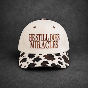 May include: A cream-colored baseball cap featuring the brown embroidered text HE STILL DOES MIRACLES on the front panel. The brim of the cap displays a brown and white cow print pattern. The cap includes small brown ventilation eyelets.