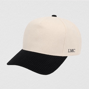 May include: A cream-colored baseball cap with a black, ribbed brim. The cap has a small black button on top and the letters "LMC" are printed on the side. The hat is a stylish accessory.