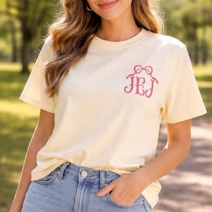 May include: A pale yellow t-shirt with a pink monogram design. The short-sleeved shirt has a crew neck and is made of a soft material. The monogram features the letters JEJ with a bow above.